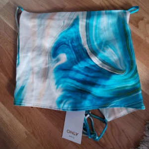 Blå satin linnetopp från ONLY - Snygg linnetopp i satin från ONLY med swirlmönster i blått, turkos, vitt och grönt. Toppen har tunna axelband och croppad passform, perfekt för en trendig look. Materialet är glansigt och mjukt mot huden. Supersnygg till jeans eller kjol!