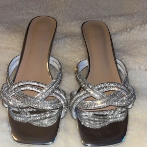 Glittriga silver sandaler med låg klack - Snygga klackar i silver med glittriga remmar täckta av strass. Skorna har fyrkantig tå, öppen tå och låg klack ca 4 cm. Perfekta för att sticka ut med en extra touch. Sula och insida i syntetmaterial. Skorna är aldrig använda, skickar direkt efter köp