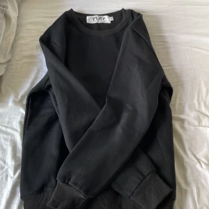 Svart sweatshirt Comme des Garçons Play - Svart sweatshirt från Comme des Garçons Play med det ikoniska hjärta-med-ögon broderiet på bröstet. Tröjan har rund hals, ribbade muddar och är tillverkad i mjuk bomull. Perfekt för dig som gillar stilrena och trendiga plagg. Använd 2 gånger, kan göra snabb affär.