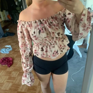 Blommig offshoulder topp från NLY One - Supersöt offshoulder topp från NLY One i ljusbeige med rosa och bruna blommor. Toppen har långa, vida ärmar och smockad resår vid axlar och midja för en snygg passform. Perfekt croppad längd och romantisk vibe.