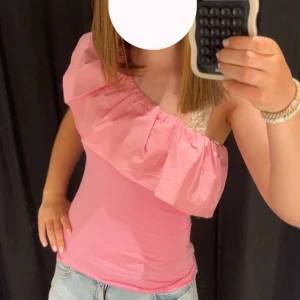 Rosa oneshoulder topp med volang - INTRESSEKOLL!! Snygg rosa topp med oneshoulder-design och stor volang längs halsringningen. Tight passform som framhäver figuren och perfekt för dig som vill sticka ut. Matcha med jeans eller kjol för en trendig look. 