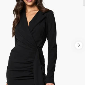 Blå omlottklänning från Boohoo - Snygg blå omlottklänning från Boohoo med lång ärm och markerad krage. Klänningen har en figurnära passform och knytdetalj i midjan som ger en cool siluett. Perfekt för dig som vill sticka ut med en djupblå färg och trendig look. Använt ca 1 gång. Köpte för 2 år sedan och  använt 1 gång sedan dess. Den finns inte längre i mörkblå färg och jag har den. Kontakta för fler bilder. Pris går att diskuteras. Passar för de i S/M