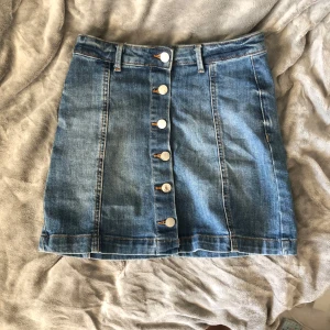 Blå jeanskjol med knappar - Snygg blå jeanskjol med silverfärgade knappar framtill. Det är två fläckar i kjolen som ni ser på siste bilden, jag tror det är fläckar från brun utan sol. Det är lite stretch i kjolen så passar nog 34-38