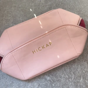 Rosa necessär från Hickap - Säljer en stilren necessär från Hickap i ljusrosa syntetmaterial med guldig dragkedja och vinrött innerfoder. Perfekt för smink eller småprylar, enkel att ta med i väskan. Oanvänd, köpt för 6 månader sen. 