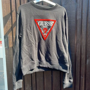 Svart sweatshirt från Guess med logga - Svart sweatshirt från Guess med stor röd och vit logga framtill. Klassisk rund halsringning och ribbade muddar vid ärmslut och nederkant. Perfekt för dig som gillar streetstyle och vill sticka ut med en ikonisk look.