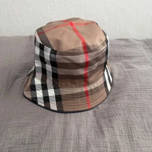 Burberry bucket hat - Snygg bucket hat från Burberry med klassiskt rutigt mönster i beige, svart, vitt och röda detaljer. Hatten har mjuk form och är tillverkad i bomull, perfekt för att lyfta din streetwear-look.