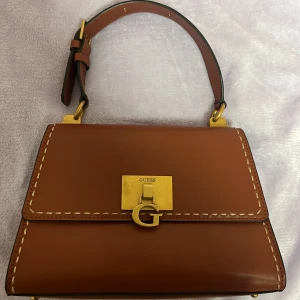 Guess väska i skinn: 2023 edition - Snygg brun handväska från Guess i slätt skinn med guldfärgade metalldetaljer och dekorativt G-spänne framtill. Väskan har justerbart handtag, kontrastsömmar och dragkedjeficka på baksidan. Insidan är ljus med Guess-logga och extra fack för småsaker. Bara använt några gånger, fortfarande i nyskick. Köpt från Zalando. Kan träffas i Stockholm om det önskas. 