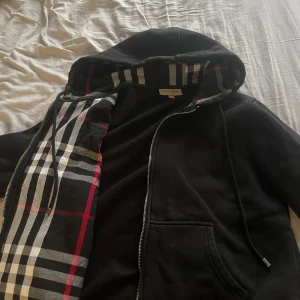 Svart hoodiejacka från Burberry  - Svart hoodiejacka från Burberry med klassiskt rutigt foder i huvan och innerkant. Jackan har dragkedja, snörning i huvan och broderad logga på bröstet. Tillverkad i mjuk bomull, perfekt för en chill och stilren look.