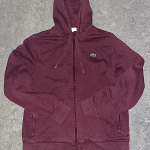 Vinröd hoodie från Lacoste med dragkedja - Snygg vinröd hoodie från Lacoste med klassisk krokodillogga på bröstet. Tröjan har huva med snörning, dragkedja framtill och två fickor. Tillverkad i mjuk bomull, perfekt för chill dagar eller när du vill ha en avslappnad stil.