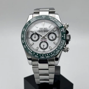 Seiko Daytona kronograf i silver - Snygg Seiko Daytona kronograf med silverfärgad länk i rostfritt stål, vit urtavla och grönt bezel. Klockan har tre svarta subdials, tydliga index och sportig känsla. Perfekt för dig som gillar stilrena accessoarer med lite edge.