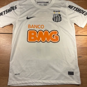 Santos Neymar Jr fotbollströja - Vit Santos FC fotbollströja från Nike med Neymar Jr och nummer 11 på ryggen. Klassisk design med svarta tryck, orange BMG-logga och klubbmärke på bröstet. Kortärmad modell i Dri-FIT-material som andas, perfekt för fotbollsplanen. Storleken är S men kan passa mindre M