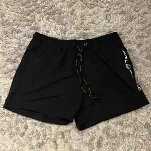 Svarta Sail Racing badshorts - Svarta badshorts från Sail Racing med vit logga längs ena benet. Shortsen har elastisk midja med snörning och en bakficka med liten logotyp. Perfekta för stranden eller poolen, tillverkade i snabbtorkande syntetmaterial.