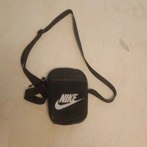 Svart axelväska från Nike - Säljer en svart axelväska från Nike med justerbar rem och dragkedja. Väskan har en vit Nike-logga på framsidan och är tillverkad i slitstark polyester. Perfekt storlek för mobil, plånbok och nycklar när du är på språng.