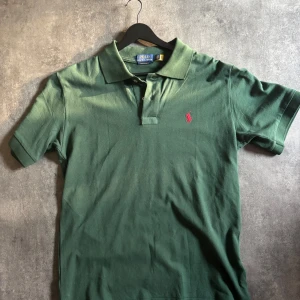 Grön pikétröja från Polo Ralph Lauren - Snygg grön pikétröja från Polo Ralph Lauren med klassisk krage och korta ärmar. Tröjan har rosa broderad logga på bröstet och är i bomull med slim fit-passform. Perfekt för en clean och stilren look.  M men passar L