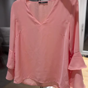 Rosa volangblus från Gina Tricot - Supersöt ljusrosa blus med v-ringning och långa ärmar med dubbla volanger. Blusen är i ett lätt, flowy material som ger en luftig känsla. 