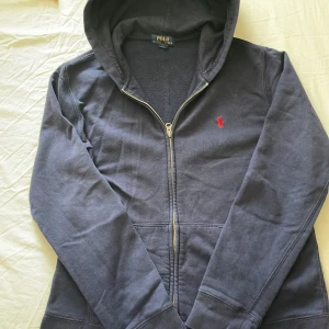Ralph lauren tröja - Mörkblå hoodie från Polo Ralph Lauren med dragkedja och huva. Klassisk röd logga broderad på bröstet och stora fickor framtill. Tillverkad i mjuk bomull, perfekt för chill dagar. Snygg och enkel att matcha med jeans eller joggers. Pris kan diskuteras😁