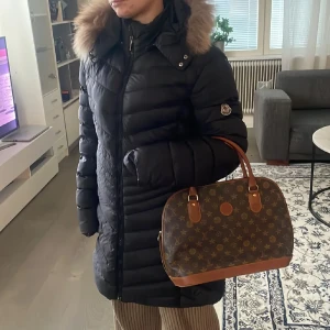 Svart Moncler dunjacka med pälskrage - Säljer en svart lång dunjacka från Moncler med snygg pälskantad huva och klassisk logga på ärmen. Jackan har dragkedja framtill, quiltad design och är perfekt för kalla dagar. Fodret har en cool serietidningsdetalj på insidan. Riktigt varm och stilren vinterjacka. Äkta såklart med qr kod se bild❤️ Nyskick