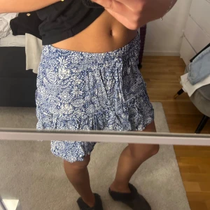 Blåvita blommiga shorts med snörning - Säljer ett par blåvita shorts med blommigt mönster och snörning i midjan. De har en lös passform och är perfekta för varma dagar. Shortsen är tillverkade i ett mjukt och lätt material som känns skönt mot huden.