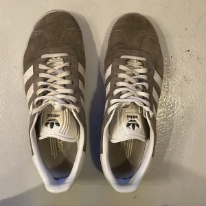 Adidas Gazelle grå sneakers - Grå Adidas Gazelle sneakers med vita ränder och vit sula. Skorna har klassisk design med snörning och rund tå. Ovandelen är i mocka och insidan är vit med Adidas-logga på hälen och plösen.