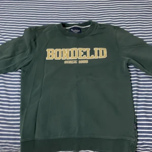 Grön sweatshirt från Bondelid - Mörkgrön sweatshirt från Bondelid med tryckt text 'BONDELID SINCE 1956' i gult och vitt på bröstet. Tröjan har rund halsringning och långa ärmar. Perfekt för en avslappnad stil.