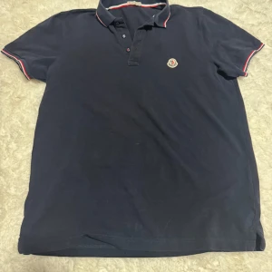 Moncler Piké - Denna klassiska Moncler pikétröja i marinblå färg är ganska använd men i bra skick. Den har en bekväm passform och passar bra i storlek, med stilrena detaljer i rött och vitt på krage och ärmslut – perfekt för en avslappnad men stilfull look.