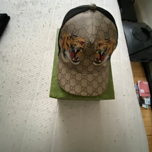 Gucci keps med tigertryck - Säljer en exklusiv Gucci keps med klassiskt GG-mönster och två stora tigrar framtill. Kepsen har svart mesh baktill och justerbar rem. Kommer med original grön Gucci-box.