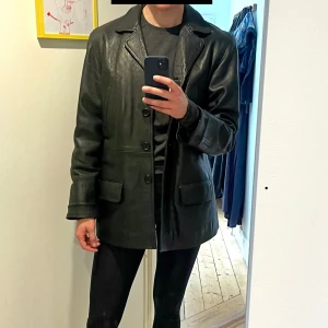 Svart skinnjacka med knappar - Säljer en svart skinnjacka med klassisk krage och knäppning framtill. Jackan har två bröstfickor med dragkedja och två fickor med lock. Snygg, rak passform och helsvart look – perfekt till många outfits. Vintage men mycket fint skick. Ärmar förlängda då de var upplagda vid köp. 