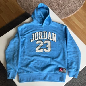 Blå Jordan hoodie med tryck - Säljer en blå hoodie från Jordan med stort vitt tryck 'JORDAN 23' på bröstet och klassisk logga nertill. Tröjan har huva och muddar vid ärmslut och nederkant. Perfekt för dig som gillar sportig stil och streetwear.