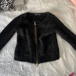Svart fluffig cardigan jacka med dragkedja - Säljer en svart, fluffig cardigan jacka med sned guldig dragkedja framtill. Jackan har lång ärm och är perfekt för dig som vill ha en mysig men stilren look.