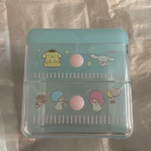 Sanrio sminkförvaringslåda med tecknade figurer - Söt sminkförvaringslåda i ljusblå plast med Sanrio-karaktärer som Pompompurin, Cinnamoroll, My Melody och Hello Kitty. Lådan har två utdragbara fack med rosa knoppar och är dekorerad med pastellfärgade motiv. Perfekt för att organisera smink eller accessoarer.