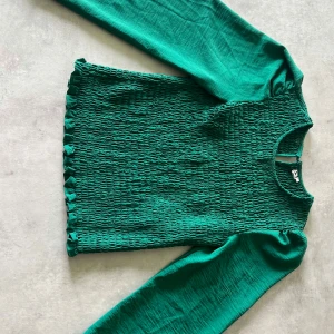 Grön smockad topp med puffärm - Säljer en grön långärmad topp med smockad kropp och puffiga ärmar. Toppen har rund halsringning och små volangdetaljer längst ner. Perfekt för dig som vill sticka ut med färg!