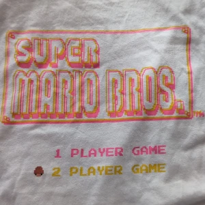Vit Super Mario Bros. t-shirt - Vit t-shirt med färgglatt Super Mario Bros.-tryck på bröstet. Klassisk passform och korta ärmar. Perfekt för dig som älskar retrospel och vill ha en cool och lekfull stil.