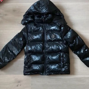 Svart glansig puffer trapstar jacka - Äkta! Originala pris:2000!Svart pufferjacka med glansig finish och huva från Philipp Plein. Jackan har dragkedja framtill, två fickor och en logotyp på bröstet. Baksidan har broderad text upptill. Perfekt för kalla dagar.