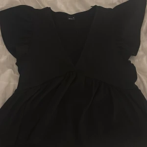 Svart blus med volangärmar - Säljer en svart blus med djup v-ringning och volangdetaljer på ärmarna. Toppen har en lös passform och markerad midja, vilket ger en fin siluett. Perfekt att styla med jeans eller kjol.