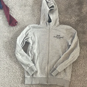 Grå hoodie från Peak Performance - Säljer en ljusgrå hoodie från Peak Performance med dragkedja och huva. Tröjan har två fickor framtill och logga på bröstet. Perfekt för en avslappnad stil och enkel att matcha med det mesta.