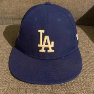 Blå LA-keps från New Era - Snygg blå keps med LA-logga framtill och New Era-logga på sidan. Klassisk modell med böjd skärm och broderade detaljer. Perfekt för dig som gillar sportig stil eller vill visa ditt stöd för Los Angeles Dodgers.