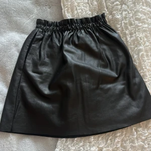 Svart kjol i skinnimitation  - Säljer en svart kortkjol i skinnimitation. Kjolen har resår i midjan för en bekväm passform och en stilren look. Perfekt att styla med en cool topp eller stickad tröja.