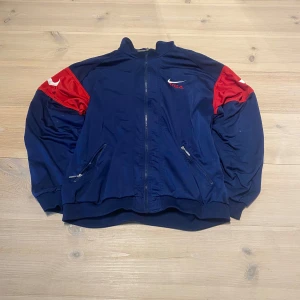 Blå och röd vindjacka från Nike - Säljer en blå jumper från Nike med röda detaljer på axlarna och vita loggor. Jackan har dragkedja framtill, två fickor med dragkedja och ribbade muddar. Snygga band med Nike-tryck längs ärmarna. Skicka ett meddelande för frågor, storlek, mått osv. 