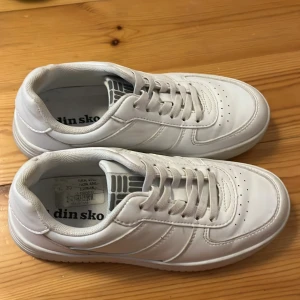 Vita sneakers från Din Sko - Säljer ett par klassiska vita sneakers från Din Sko med rund tå och platt sula. Skorna har snörning och är tillverkade i syntetmaterial. Perfekta för en clean och stilren look.
