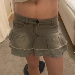 Grå volangkjol i jeans - Säljer en grå kort volangkjol i jeansmaterial med två volanger och klassiska jeansdetaljer som fickor och knapp framtill. Perfekt för dig som gillar en avslappnad men trendig stil. PASSAR MIG MED S