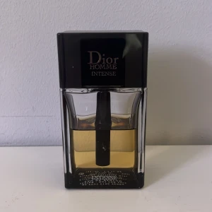Dior Homme Intense Eau de Parfum - Dior Homme Intense är en lyxig herrparfym med en elegant och maskulin doft. Flaskan är rektangulär i genomskinligt glas med svart lock och detaljer. Innehållet är gyllene och flaskan har en modern, stilren design.