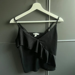 Svart croppad linnetopp med volang - Säljer en svart croppad linnetopp med smala axelband och volangdetalj framtill. Perfekt för varma dagar eller att styla lager på lager. Enkel och stilren design.