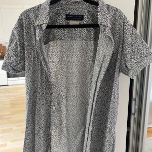 Vit blommig kortärmad skjorta från Jack & Jones - Säljer en vit kortärmad skjorta från Jack & Jones med små mörkblå blommor över hela tyget. Skjortan har klassisk krage och knäppning framtill. Perfekt för en fräsch och stilren look i sommar.