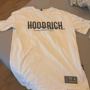 Vit t-shirt från Hoodrich med tryck - Vit t-shirt från Hoodrich med blått tryck framtill och mindre tryck på ryggen. Klassisk passform med rund hals och korta ärmar. Märkesdetaljer nertill på framsidan. Perfekt för en avslappnad streetwear-look.