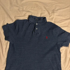 Blå pikétröja från Polo Ralph Lauren - Säljer en blå pikétröja från Polo Ralph Lauren i slim fit-modell. Tröjan har korta ärmar, klassisk krage och två knappar framtill. Den är dekorerad med det ikoniska röda broderade logotypen på bröstet.
