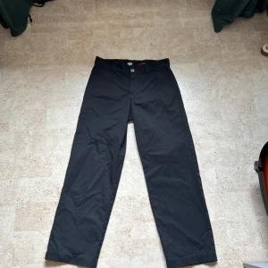 Svarta kostymbyxor från Dickies storlek W30 L32 - Säljer ett par svarta kostymbyxor från Dickies med klassisk passform och raka ben. Byxorna har dragkedja och knapp framtill samt Dickies-logga ovanför ena bakfickan. Perfekta för en clean och stilren look.