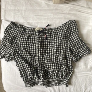Svartvit rutig offshoulder blus - Säljer denna jättesöta blus med rutigt mönster och offshoulder- modell. Blusen har resår vid axlar och midja samt en liten rosett framtill. I storlek 158 (ungefär xs/xxs). Använd endast några gånger så i bra skick!