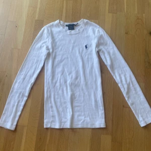 Vit långärmad tröja från Ralph Lauren - Säljer en klassisk vit långärmad tröja från Ralph Lauren i 100% bomull. Tröjan har en tight passform och pryds av den ikoniska blå loggan broderad på bröstet. Perfekt basplagg till garderoben!