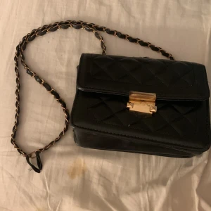 Svart quiltad axelväska med gulddetaljer - Säljer en svart quiltad axelväska med kedjerem och snyggt guldfärgat spänne framtill. Väskan har klassisk design och är perfekt för att lyfta din outfit. Liten och smidig storlek, passar när du vill bära det viktigaste.