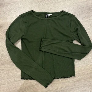 Mörkgrön ribbad långärmad topp från H&M - Säljer en mörkgrön ribbad långärmad topp från H&m. Toppen har croppad passform och vågig kant nertill för en trendig look. Perfekt att styla med jeans eller kjol.